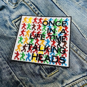 May include: Square patch with the text "ONCE IN A LIFETIME TALKING HEADS" in black, with colourful figures. The patch is on a denim jacket.