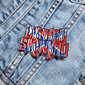Può includere: Una toppa ricamata rosso, bianco e blu con il testo "Lynyrd Skynyrd" in nero. La toppa è cucita su una giacca di jeans blu.