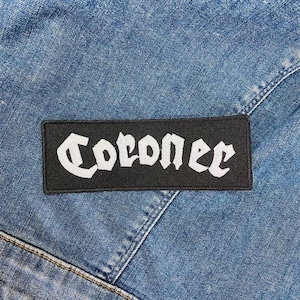 Peut inclure: Un écusson rectangulaire noir avec le mot "Coroner" en lettres gothiques blanches. L'écusson est posé sur un fond en denim bleu, avec des coutures visibles.