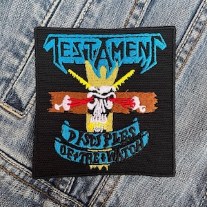 May include: A black embroidered patch with the band name "TESTAMENT" in blue and yellow lettering. The patch features a skull with red eyes and a cross with a brown background. The text "DISCIPLES OF THE WATCH" is written in blue and yellow lettering on a blue banner.
