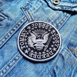 May include: A black and white embroidered patch with the text "Johnny Depp" and an eagle design. The patch is on a blue denim jacket.