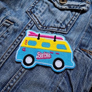 Peut inclure: Un patch brodé bleu et jaune représentant un van vintage avec le mot "Barbie" sur le côté. Le van a une planche de surf sur le toit et des roues blanches.
