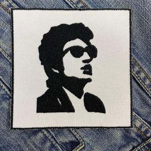 May include: Black and white embroidered patch of a person wearing sunglasses. The person has short, dark hair and is wearing a jacket.