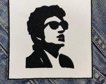 May include: Black and white embroidered patch of a person wearing sunglasses. The person has short, dark hair and is wearing a jacket.