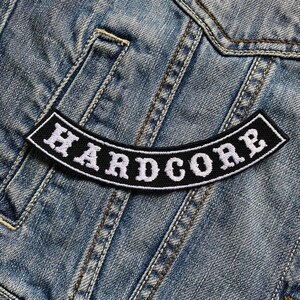 Può includere: Toppa ricamata in bianco e nero con la parola "HARDCORE" in lettere bianche su sfondo nero. La toppa è cucita su una giacca di jeans blu.