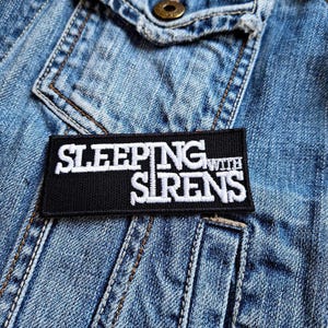 May include: Black and white embroidered patch with the text "Sleeping With Sirens" on a denim jacket.