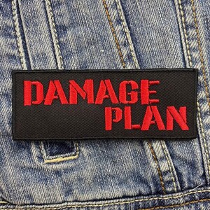 Puede incluir: Parche negro con texto bordado en rojo que dice "DAMAGE PLAN".