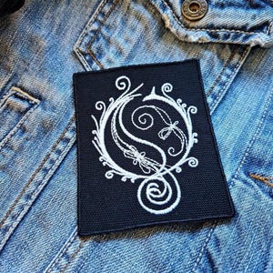May include: A black embroidered patch featuring a white, ornate design. The design is a stylized monogram or emblem with swirling lines and decorative elements. The patch is rectangular and is placed on a denim jacket.