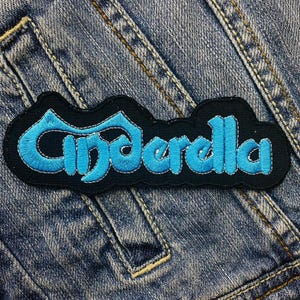 Può includere: Una toppa ricamata nera e blu con la parola "Cenerentola" in un font stilizzato.