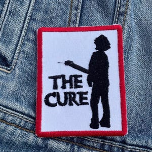 May include: A white patch with a red border featuring a black silhouette of a person holding a microphone and the text "THE CURE" in black.