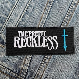 Op de afbeelding: Zwarte en witte geborduurde patch met de tekst "The Pretty Reckless" en een gestileerd blauw kruis.