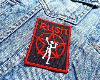 Peut inclure: Un écusson brodé noir et rouge avec le mot "Rush" et une étoile stylisée avec une figure à l'intérieur. L'écusson est cousu sur une veste en jean bleue.