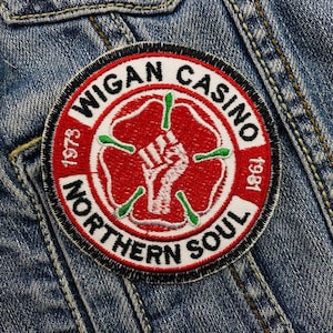 Puede incluir: Un parche bordado en blanco y negro con una rosa roja y un puño en alto. El texto alrededor de la rosa dice "Wigan Casino 1973 Northern Soul 1981".