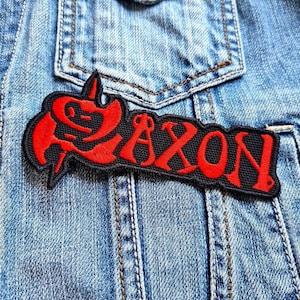 May include: Red and black embroidered patch with the word "SAXON" in a stylized font. The patch is on a blue denim jacket.