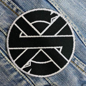 Puede incluir: Parche bordado en blanco y negro con un diseño circular. El diseño presenta una serie de líneas y formas que se cruzan.