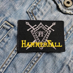 May include: Black embroidered patch with a white shield and two crossed swords. The shield has the letters "HF" inside. The text "HAMMERFALL" is embroidered in yellow below the shield.