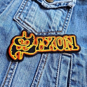 Könnte beinhalten: Ein schwarzer und gelber gestickter Aufnäher mit dem Wort "SAXON" in einer stilisierten Schriftart. Der Aufnäher befindet sich auf einer blauen Jeansjacke.