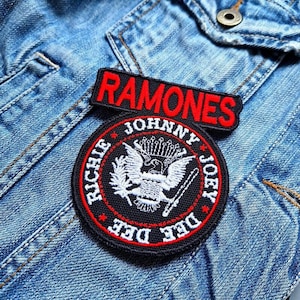 Könnte beinhalten: Ein gestickter schwarzer und weißer Aufnäher mit den Worten "Ramones", "Johnny", "Joey", "Richie" und "Dee Dee" in einem Kreis. Der Aufnäher befindet sich auf einer blauen Jeansjacke.