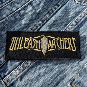 May include: Black patch with white stitching featuring the text "UNLEASH THE ARCHERS" in gold lettering with a stylized silver sword design.