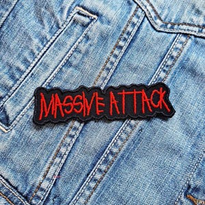 Könnte beinhalten: Ein schwarzer Aufnäher mit den roten Worten "MASSIVE ATTACK", auf eine hellblaue Jeansjacke genäht. Der Aufnäher hat ein gezacktes Kantendesign.