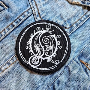 May include: A black and white embroidered patch with a stylized letter "C" surrounded by intricate floral designs.