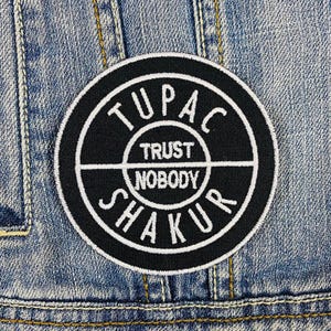 Puede incluir: Parche bordado en blanco y negro con el texto "Tupac Trust Nobody Shakur" en un círculo.