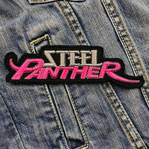 Peut inclure: Un patch brodé noir et rose avec le texte "Steel Panther" dans une police de caractères stylisée.