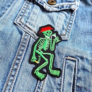 Peut inclure: Squelette vert portant un chapeau rouge et tenant un verre de liquide. Le squelette est assis sur une veste en jean.