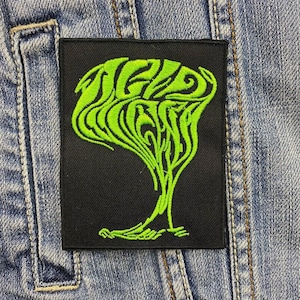 May include: Green embroidered patch with a swirling design on a black background. The design resembles a tree or a plant.