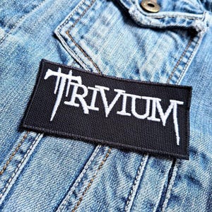 Peut inclure: Un écusson rectangulaire noir avec le mot "TRIVIUM" brodé en blanc, posé sur une veste en jean bleu clair. L'écusson présente une police de caractères stylisée. La veste en jean montre des coutures visibles.