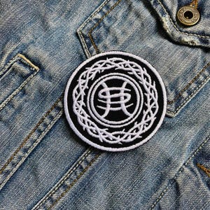 Puede incluir: Un parche redondo bordado en blanco y negro. El parche presenta un diseño blanco estilizado de una corona de espinas alrededor de un emblema central. El parche está adherido a una chaqueta vaquera azul claro.