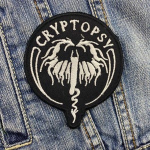 May include: Black and white embroidered patch with the text "CRYPTOPSY" in a circle around a white, stylized, dragon-like creature.