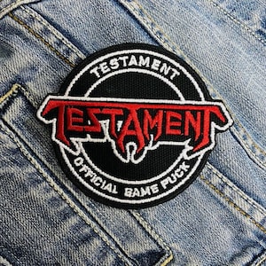 May include: A black and white circular patch with the band name "Testament" in red lettering. The patch also says "Official Game Puck" in white lettering.