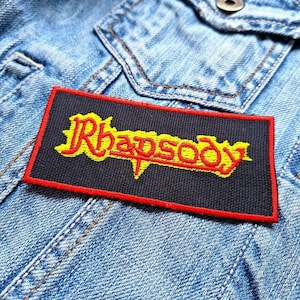 Op de afbeelding: Een zwarte en rode geborduurde patch met het woord "Rhapsody" in gele letters.