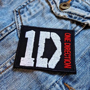 May include: A black rectangular patch embroidered with the white letters "1D" and the red text "ONE DIRECTION". The patch is on a denim jacket. The patch is rectangular and has a black background.