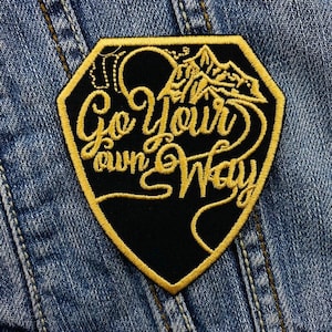 Op de afbeelding: Een geborduurde zwarte en gouden patch met de tekst "Go Your Own Way" in een cursief schrift.