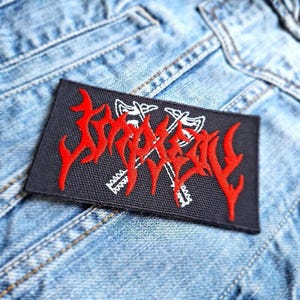 Könnte beinhalten: Roter gestickter Patch mit dem Wort "SLAUGHTER" in einer stilisierten Schriftart, mit zwei gekreuzten Äxten in Weiß hinter dem Text. Der Patch befindet sich auf einem dunkelblauen Jeansstoff.