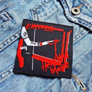 Puede incluir: Parche bordado en negro y rojo con el texto "Exciter" y "Heavy Metal" en una fuente estilizada. El parche presenta una figura roja y blanca sobre un fondo rojo.