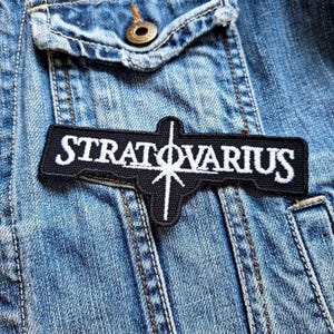 Puede incluir: Un parche bordado negro con la palabra "STRATOVARIUS" en letras blancas. El parche tiene una forma única con un diseño en forma de cruz y un elemento de estrella. Está fijado a una chaqueta vaquera.