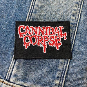 Może przedstawiać: Czarno-biała naszywka haftowana z napisem "Cannibal Corpse" w czerwonych literach. Naszywka znajduje się na dżinsowym tle.