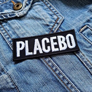 Puede incluir: Un parche negro con costuras blancas que dice "PLACEBO" en una chaqueta vaquera azul.