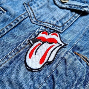 May include: A white and red embroidered patch of a tongue sticking out with a black outline. The patch is sewn onto a blue denim jacket.