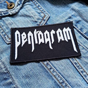 Op de afbeelding: Een zwarte patch met witte tekst die "PENTAGRAM" zegt, genaaid op een blauw denim jack.
