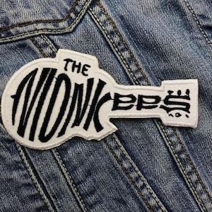 May include: White embroidered patch with black lettering that reads "The Monkees" on a denim background.