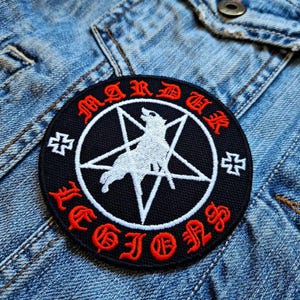 Può includere: Una toppa rotonda nera con testo rosso e bianco e un lupo bianco all'interno di un pentagramma. Il testo recita "MARDUK LEGIONS" in un carattere gotico. Due croci bianche sono ai lati. La toppa è su una giacca di jeans.