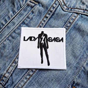 Peut inclure: Un écusson carré blanc avec les mots "LADY GAGA" en noir, avec une silhouette noire d'une personne et un éclair. L'écusson est posé sur une veste en jean bleue.