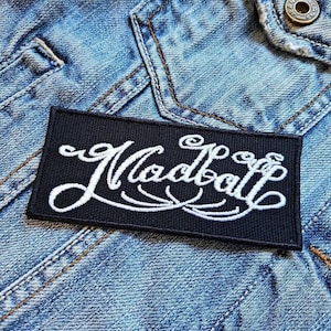 May include: A rectangular black patch with the word "Madball" embroidered in white script. The patch is placed on a light blue denim jacket, with visible stitching and a button.