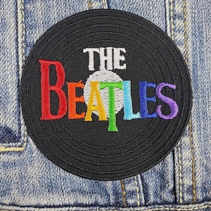 Peut inclure: Un patch brodé noir et blanc représentant un disque vinyle avec le texte "THE BEATLES" en couleurs arc-en-ciel.