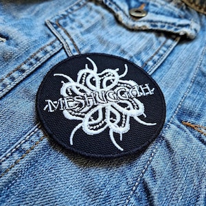 Op de afbeelding: Een zwart-witte geborduurde patch met de bandnaam "Meshuggah" in witte letters. Het ontwerp heeft een gestileerd, abstract, wit bloemenpatroon.