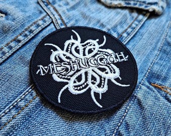 Może przedstawiać: Naszywka haftowana w kolorze czarnym i białym z nazwą zespołu "Meshuggah" w białych literach. Projekt zawiera stylizowany, abstrakcyjny, biały wzór kwiatowy.